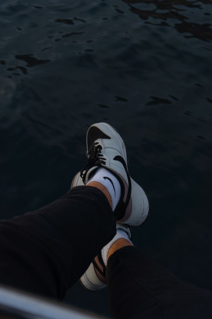 Home Person relaxing by the waterfront with legs crossed, showcasing trendy sneakers against a dark water backdrop.
